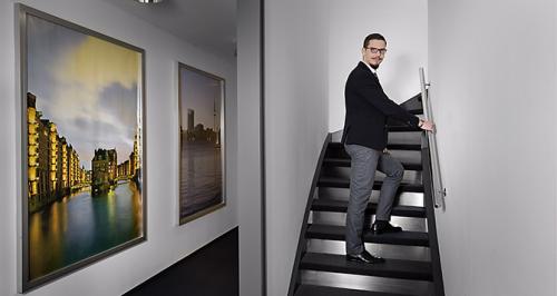 Mitarbeiterfoto eines Mannes in einem Treppenhaus mit Hamburger Bildmotiven an den Wänden.